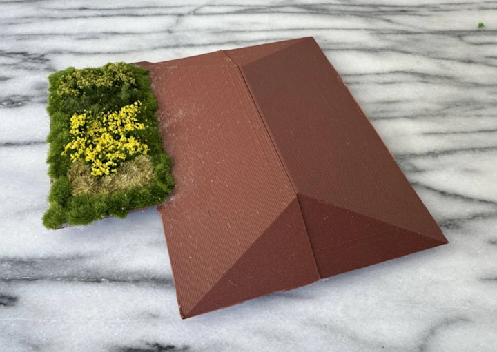 A close-up of two 3D-printed roof sections resting on a marble surface. One section is a brown gabled roof, and the other is a green roof topped with textured model grass and small yellow plants, representing sustainable landscaping in an architectural model.